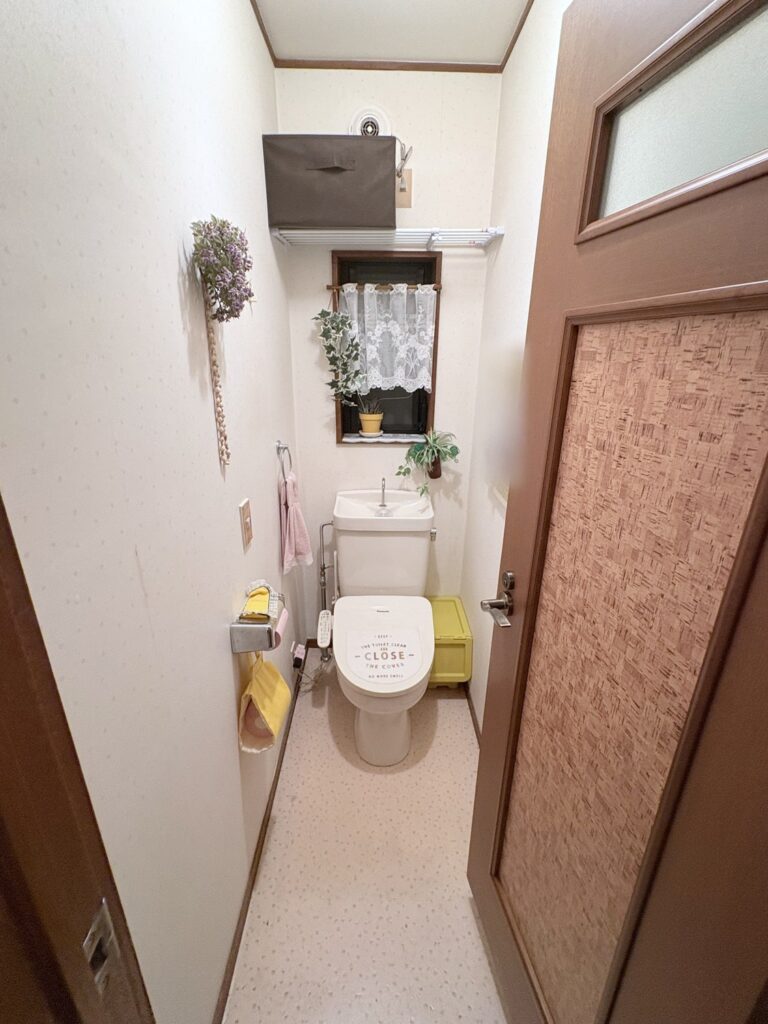 トイレのBefore。白い便器に「CLOSE」と書かれたふたのカバー。上の棚に黒い収納ボックス、左の壁にラベンダー色の壁飾り、便器のまわりに黄色の小物があり、右にコルク柄のドアがある