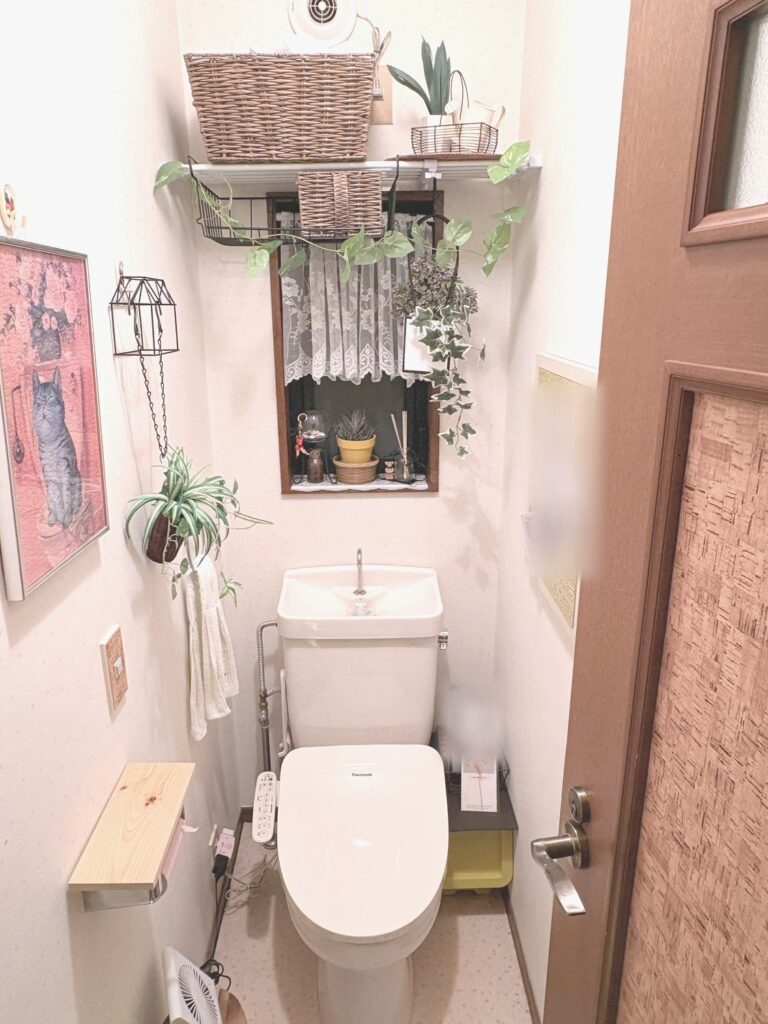 飾りつけたトイレを正面から見たところ。上の棚にラタン風のかご大小、窓まわりにレースのカーテンと吊るした植物、左の壁に猫のジグソーパズルのアート、家型のワイヤーの飾りと吊るした植物、白いフェイスタオル
