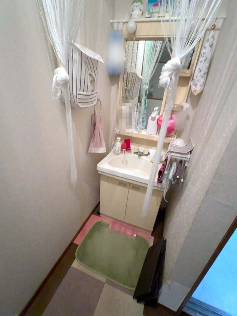 飾る前の2階の洗面所。白い紐カーテン、ストライプの吊り下げポーチ、ピンクのタオル、ピンクのじょうろ、緑とピンクのマット、鏡の上の棚に夏の飾り