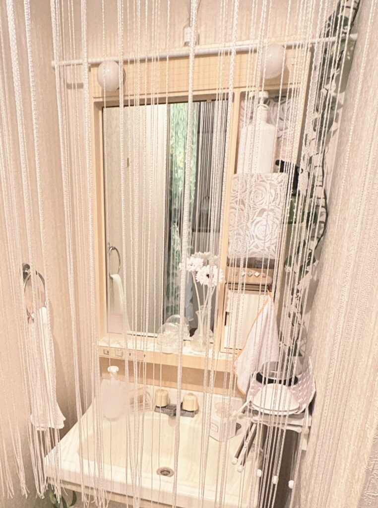 紐カーテン越しに見た、夜の洗面台の鏡のまわり。鏡の上に丸い電球が2つ、鏡の前に白いガーベラ、右にグリーン