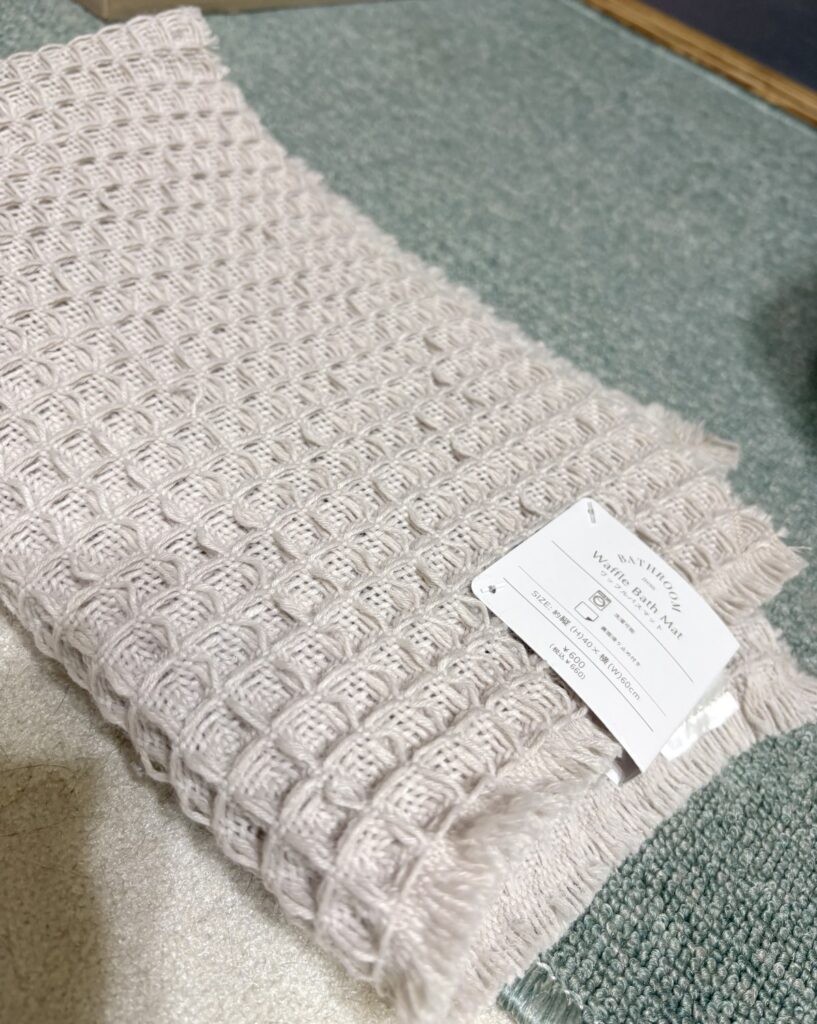 3COINSのワッフル織りのバスマット。うすいベージュで、ふちにフリンジ。タグに「Waffle Bath Mat」「約縦40×横60cm」「税込¥660」
