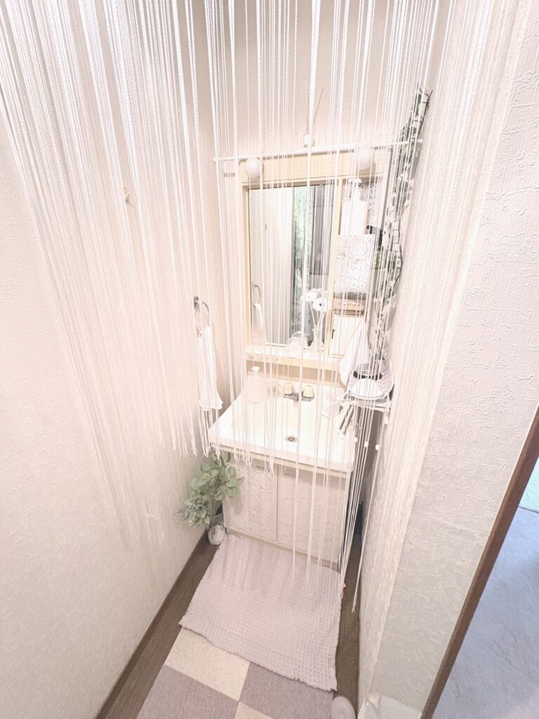 白い紐カーテン越しに見た今の洗面所。洗面台の前にワッフル織りのマット、左の床にグリーンを挿した花瓶、鏡のまわりに白いガーベラ、右にグリーン