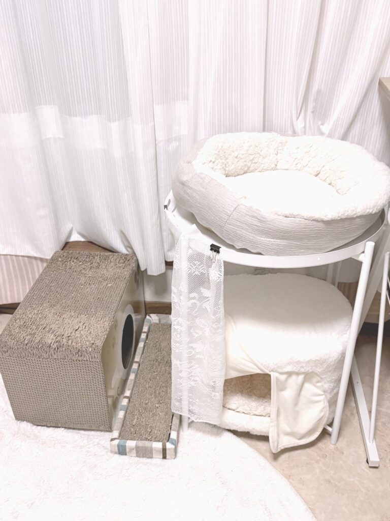 GLADOMのテーブルの下に、白いふわふわの屋根付きハウスを置いたところ。上には丸いペットベッド、左には段ボールの爪とぎが2つある
